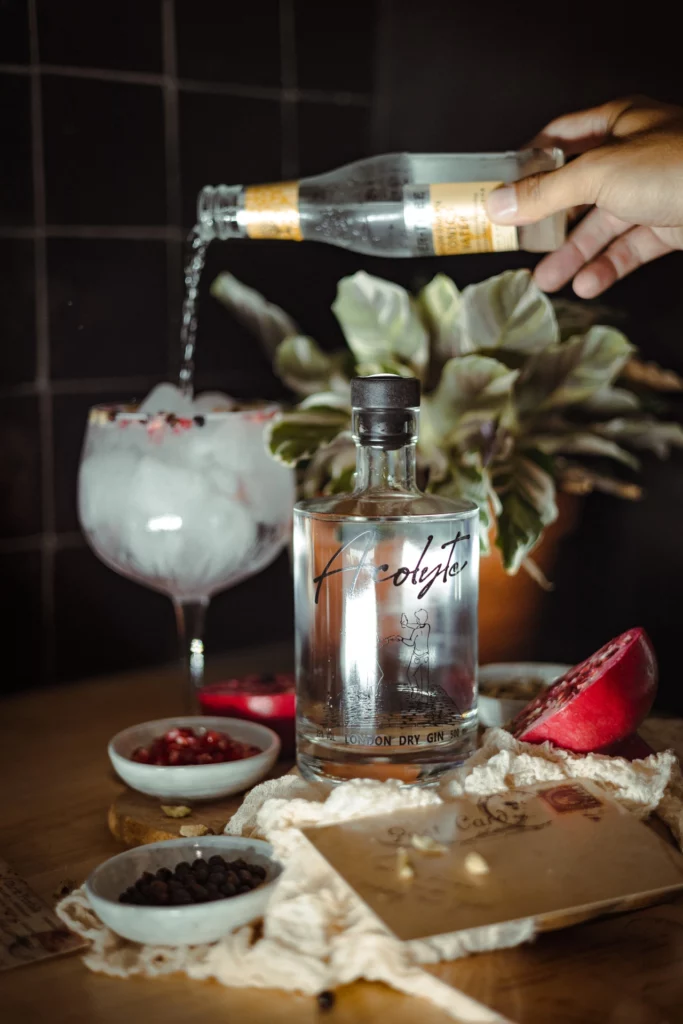 Une bouteille de gin Acolyte entourée de ces saveurs les plus importantes . Et la préparation d'un cocktail avec ce gin et un tonic en arrière-plan.