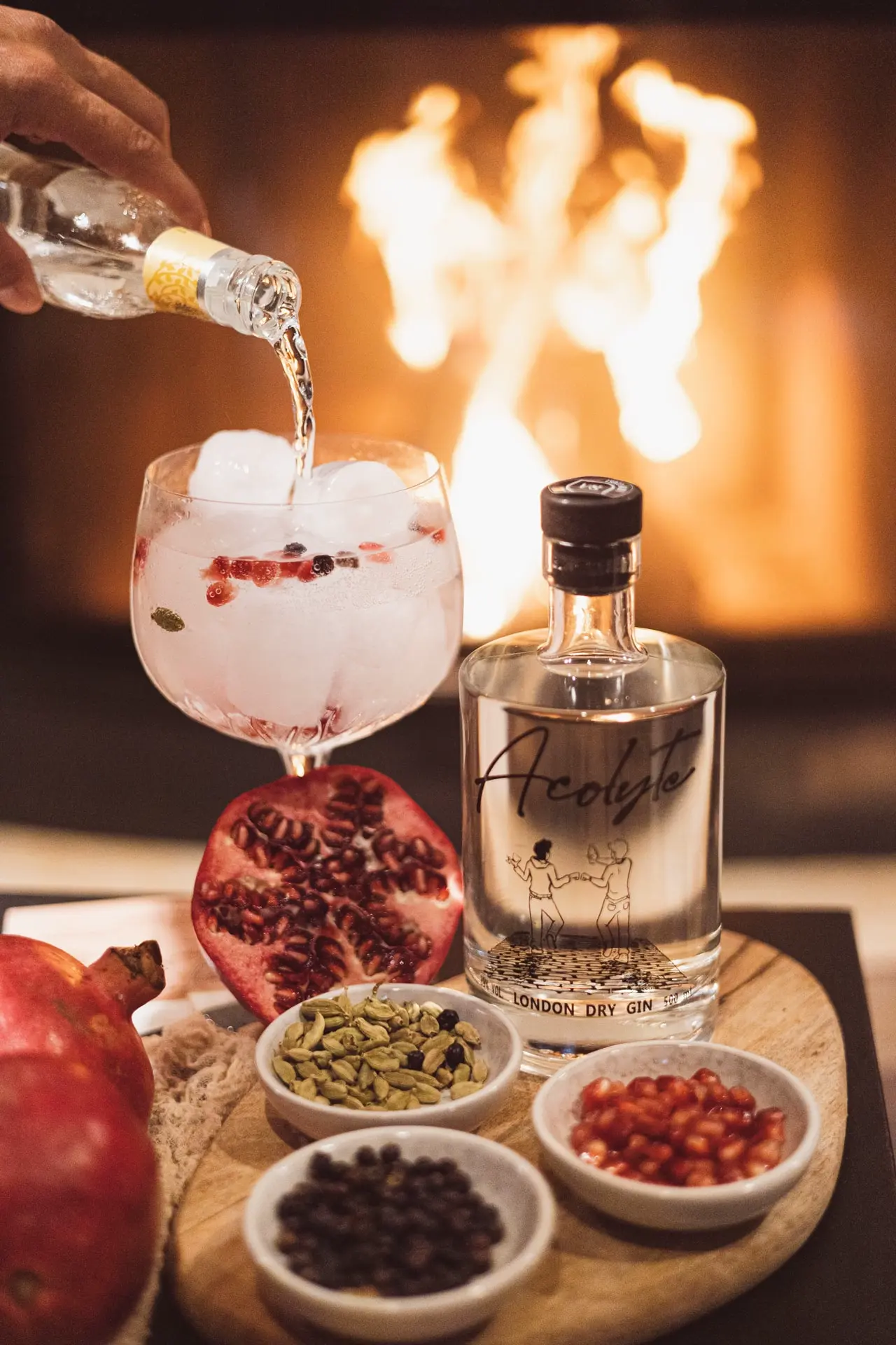 Bouteille de Gin Acolyte entourée de ses saveurs principale. Avec un feu en fond ainsi qu'un verre avec un gin en préparation.